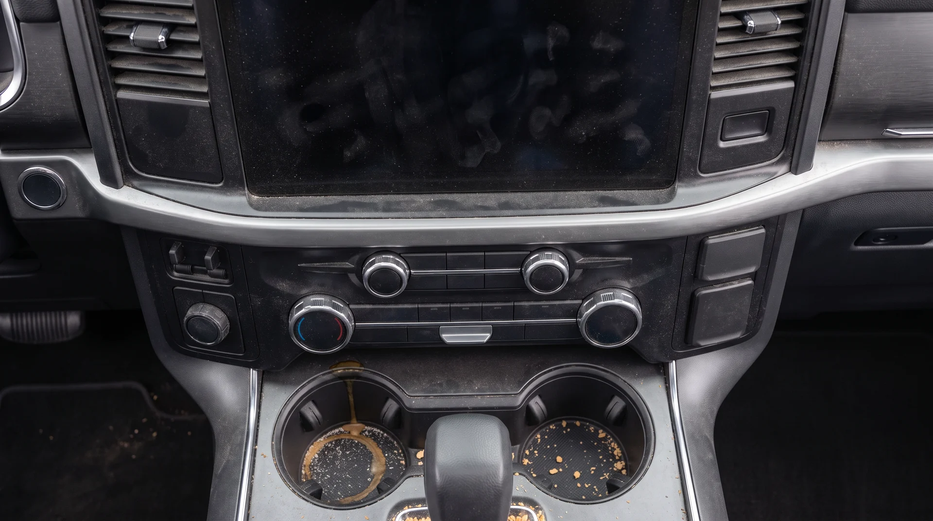 A center console before detailing, dust across the screen and vents and crumbs and a dried coffee ring in the cupholders